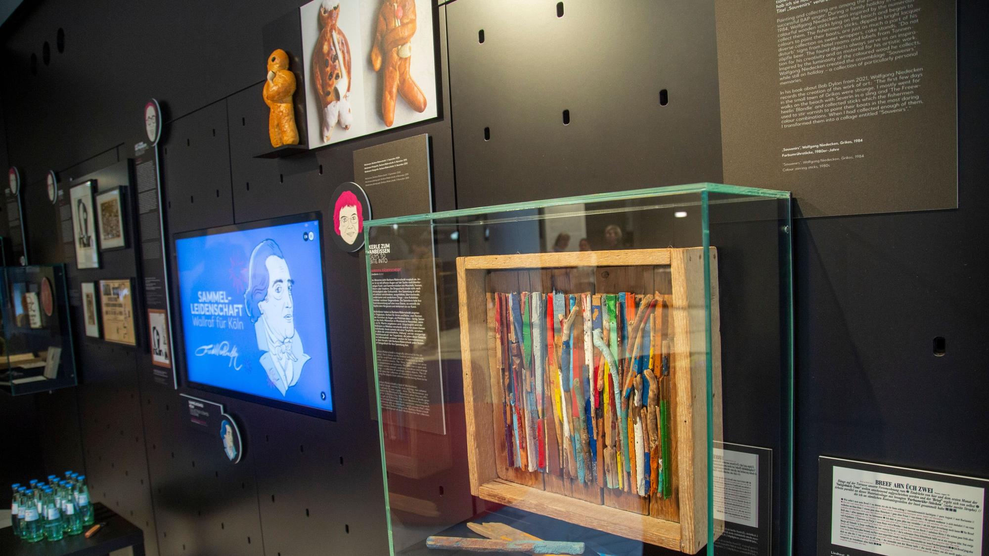 Blick in den Ausstellungsraum: Die bunten Holzstäbe im Vordergrund hat BAP-Sänger Wolfgang Niedecken im Familienurlaub 1984 am Strand von Griechenland gesammelt und zu einer Assemblage verarbeitet. Im Hintergrund der Bildschirm mit Ferdinand Franz Wallraf und ein Foto von Weckmännern.