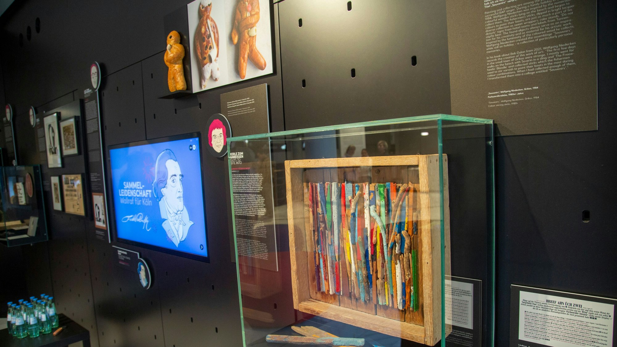 Blick in den Ausstellungsraum: Die bunten Holzstäbe im Vordergrund hat BAP-Sänger Wolfgang Niedecken im Familienurlaub 1984 am Strand von Griechenland gesammelt und zu einer Assemblage verarbeitet. Im Hintergrund der Bildschirm mit Ferdinand Franz Wallraf und ein Foto von Weckmännern.