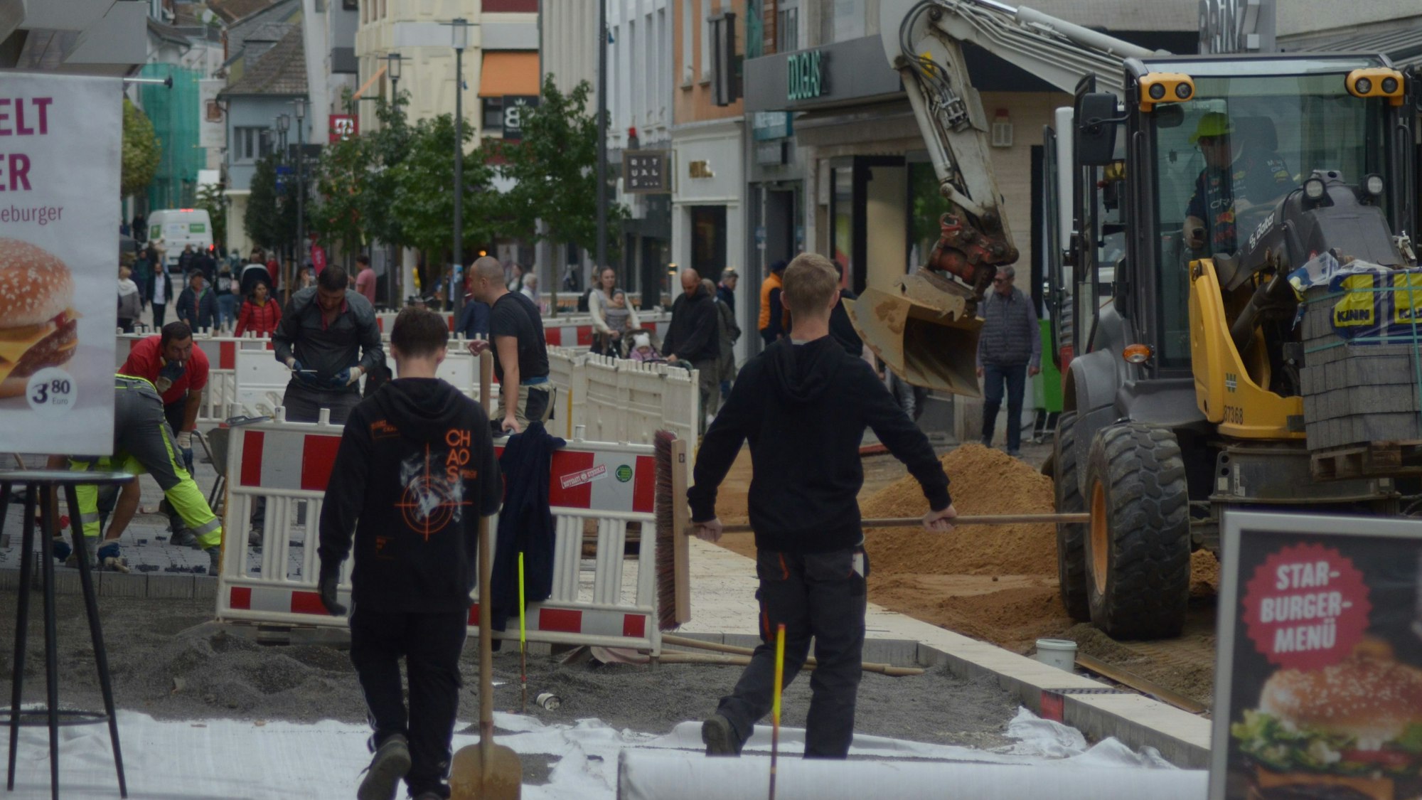 In der Neustraße verlegen Arbeiter hinter Absperrzäunen Pflastersteine. Ein Bagger verteilt Sand, während Passanten sich einen Weg vorbei an der Baustelle bahnen.