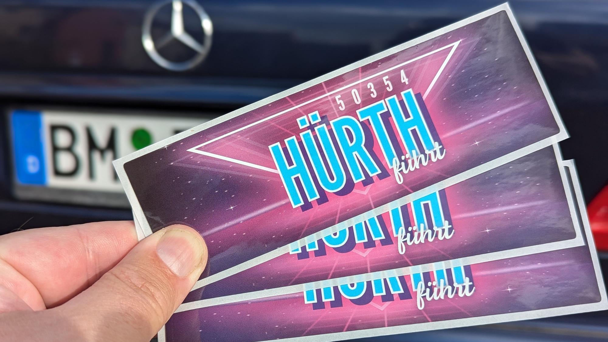 Das Foto zeigt eine Hand, die drei Aufkleber vor einem Auto mit BM-Kennzeichen hält.