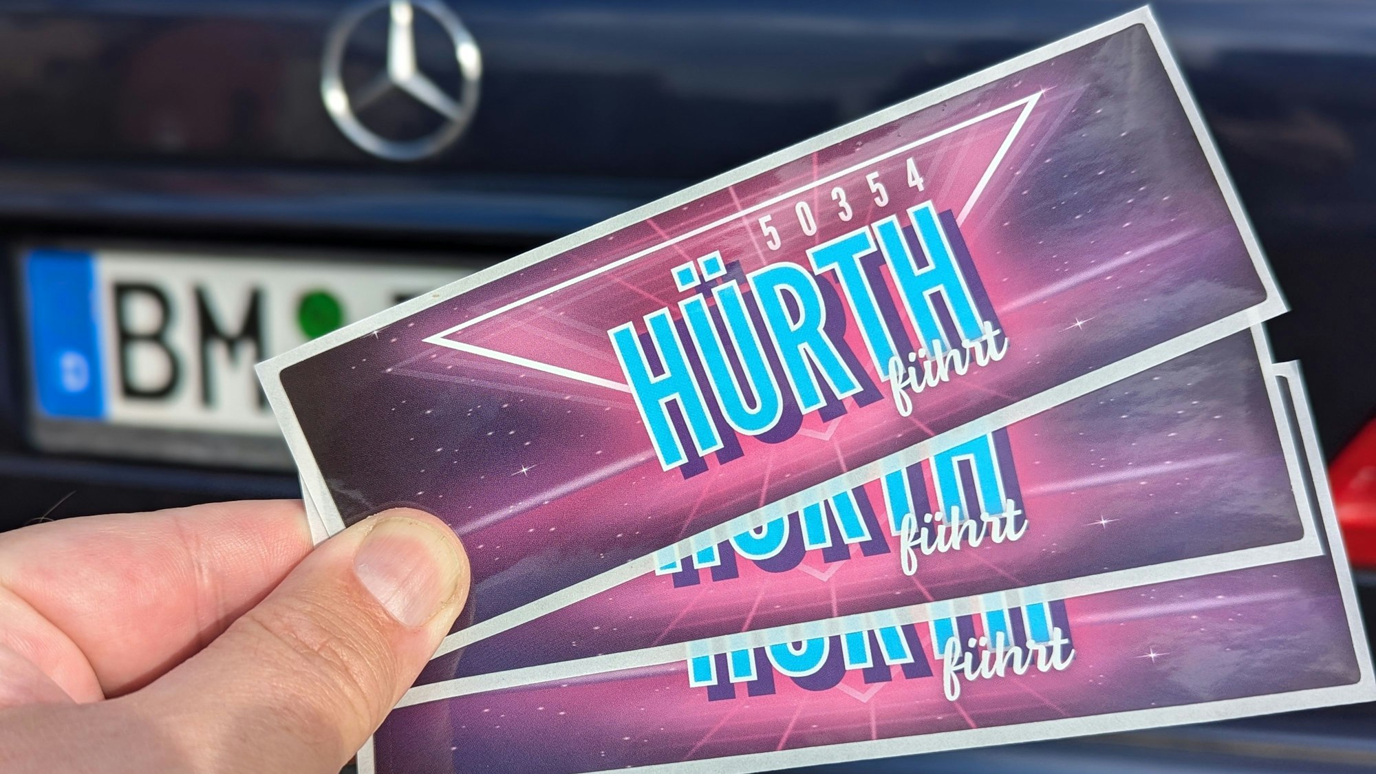 Das Foto zeigt eine Hand, die drei Aufkleber vor einem Auto mit BM-Kennzeichen hält.