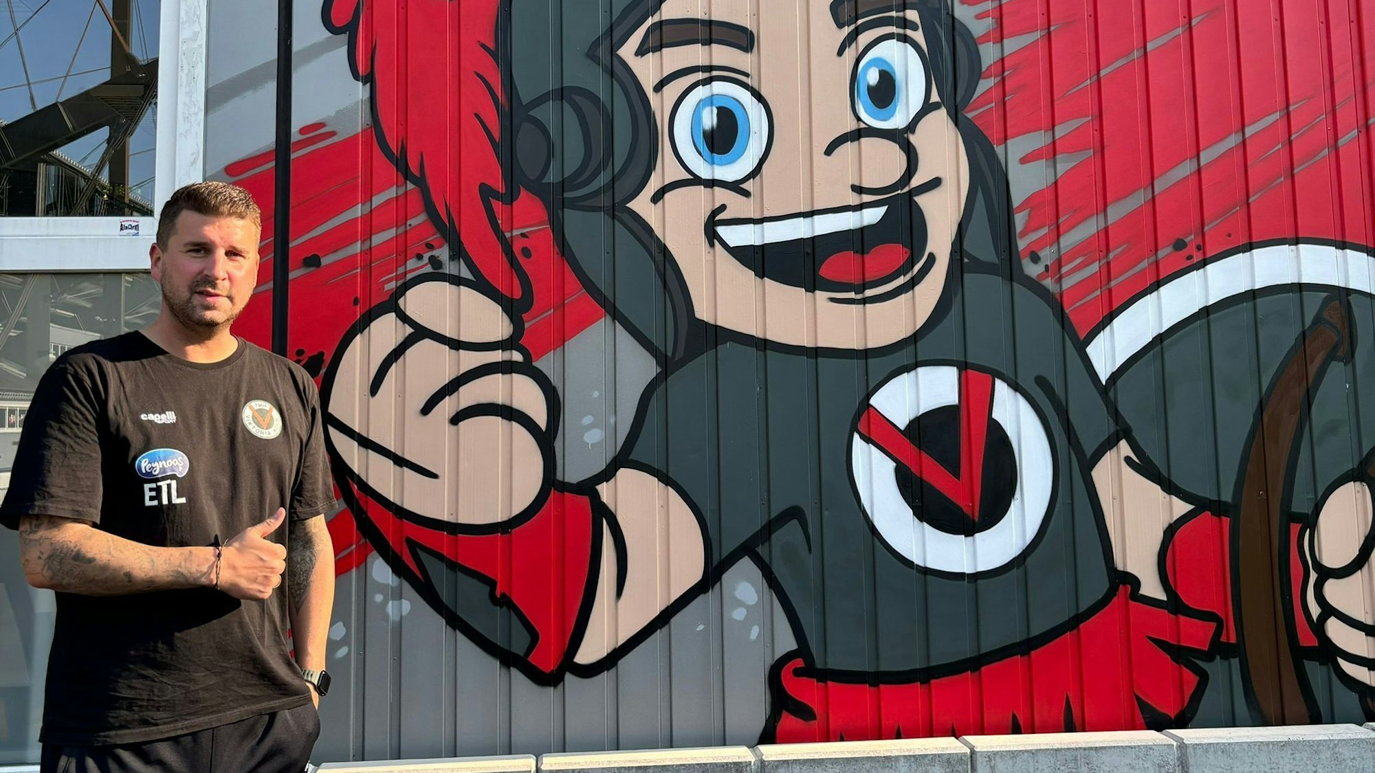 Christian Hammes steht vor einer Wand, auf die das Maskottchen von Viktoria Köln gezeichnet wurde.