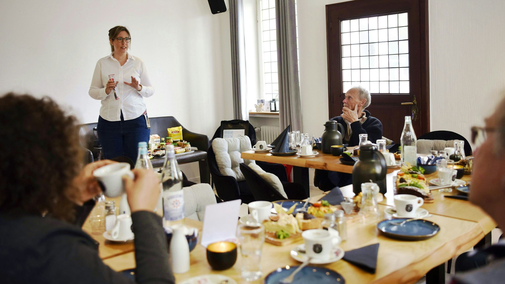 Angelika Eimermacher vom DRK Euskirchen steht in einer weißen Bluse und dunkler Jeans vor einer Gruppe Menschen, die an Tischen sitzen und Kaffee trinken. Auf den Tischen stehen Kaffeetassen, Kuchen und weiteres Geschirr.