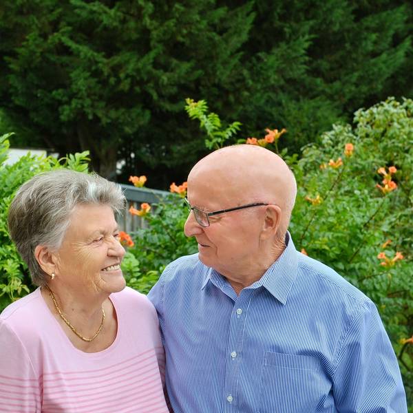 Anna und Heinrich Wagner stehen vor grünen Büschen und schauen sich an. Anna Wagner trägt ein rosafarbenes Oberteil, eine goldene Kette, kleine goldene Ohrringe und kurze graue Haare. Heinrich Wagner trägt ein blaues Hemd und eine Brille, er hat eine Glatze.