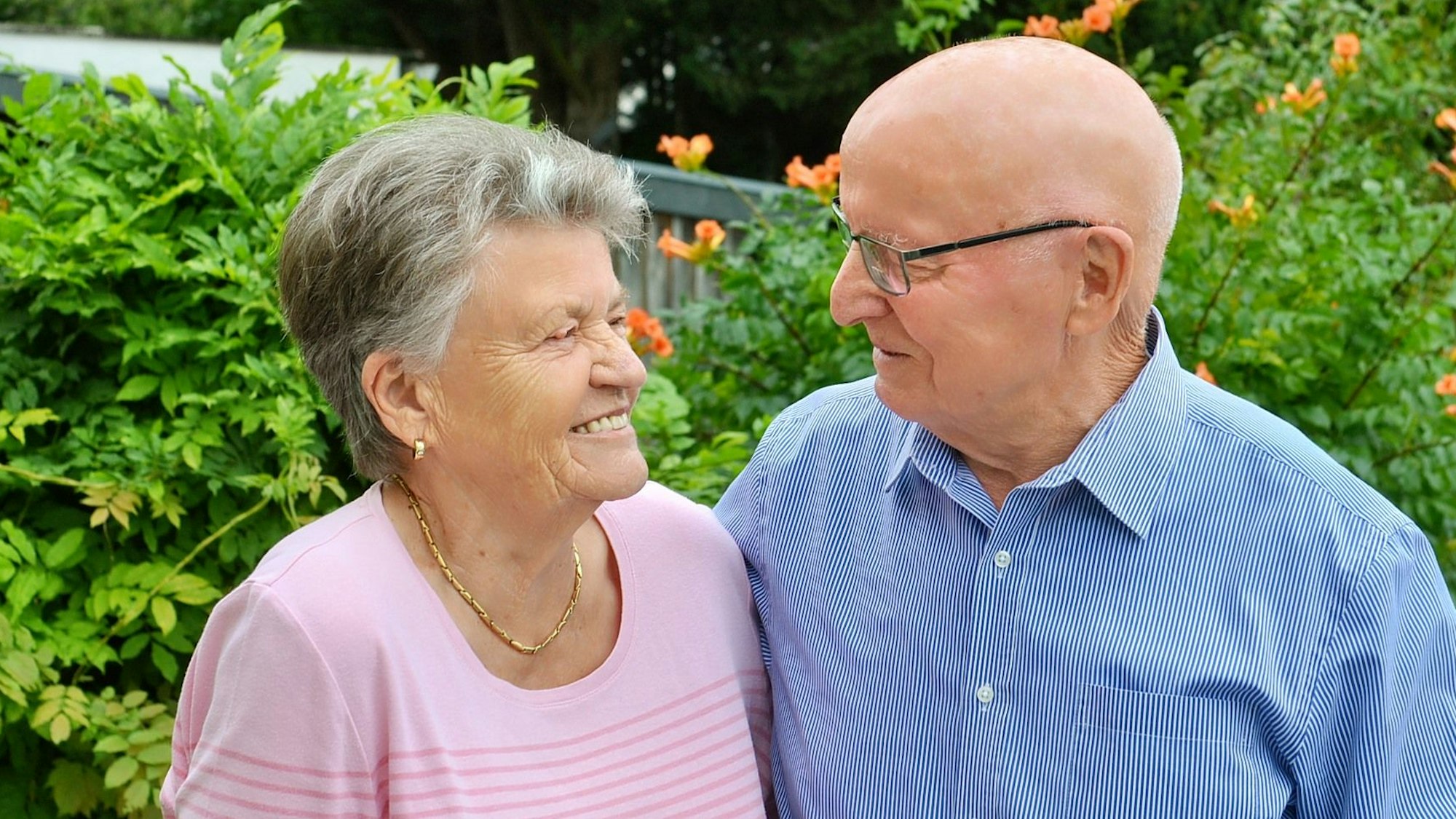 Anna und Heinrich Wagner stehen vor grünen Büschen und schauen sich an. Anna Wagner trägt ein rosafarbenes Oberteil, eine goldene Kette, kleine goldene Ohrringe und kurze graue Haare. Heinrich Wagner trägt ein blaues Hemd und eine Brille, er hat eine Glatze.