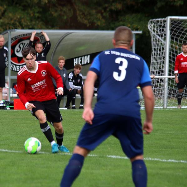 Ein Fußballer hat den Ball am Fuß und setzt zum Dribbling an. Im Vordergrund beobachtet ihn ein Verteidiger.