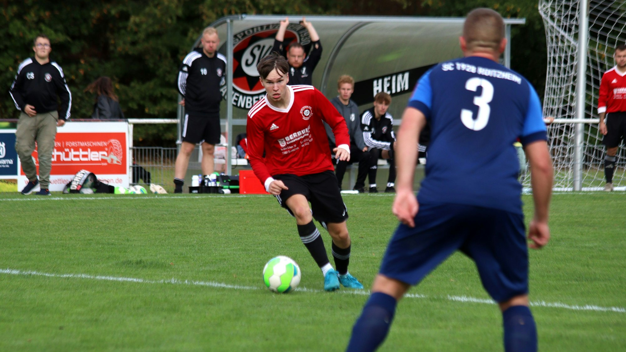 Ein Fußballer hat den Ball am Fuß und setzt zum Dribbling an. Im Vordergrund beobachtet ihn ein Verteidiger.