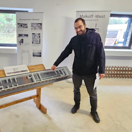 Historiker Julian Weller steht in dunkler Regenjacke und dunkler Jeans neben einem Modell der neuen Ausstellung in Vogelsang. Hinter ihm sind Fenster und Aufsteller zu sehen.