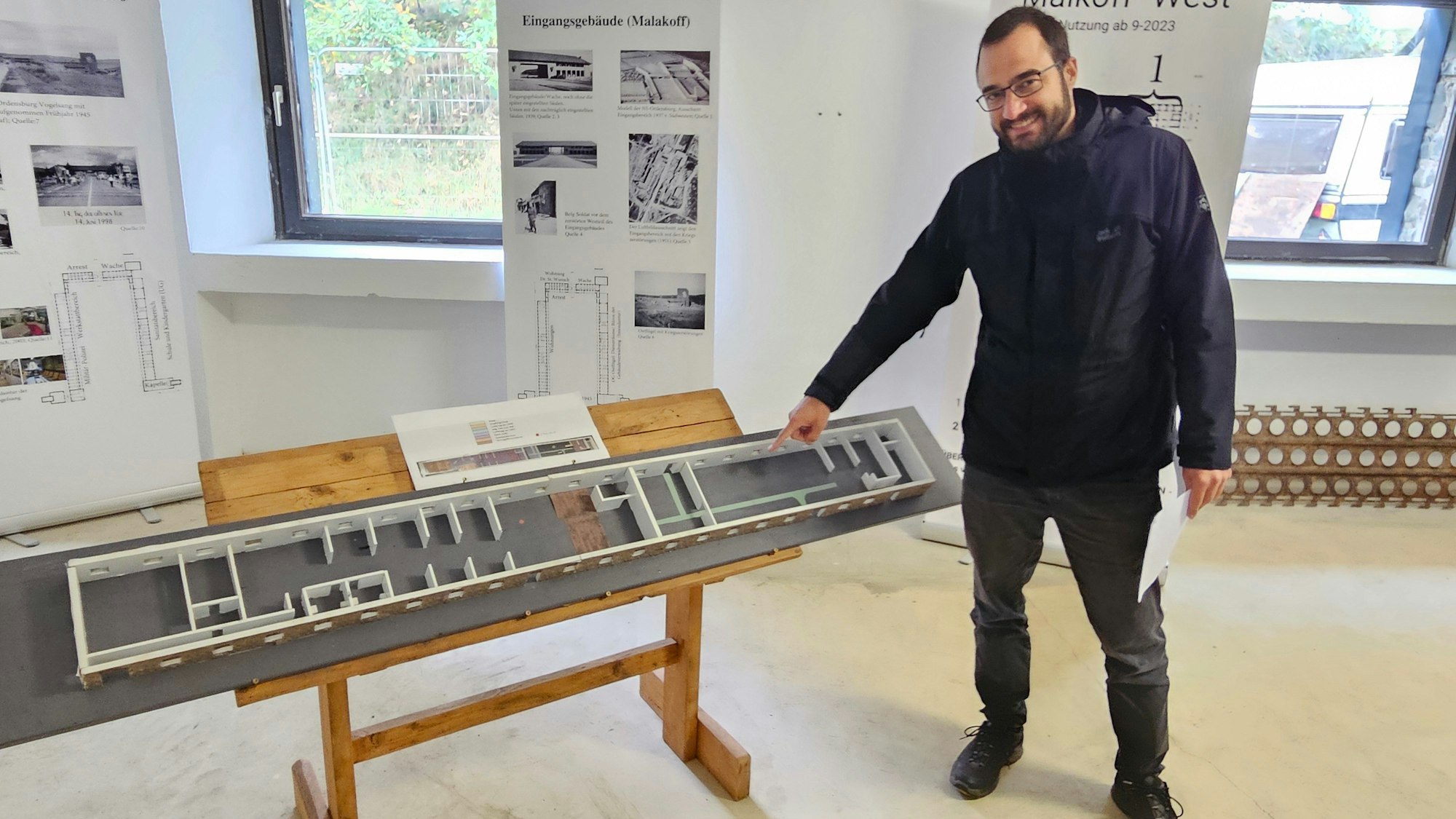 Historiker Julian Weller steht in dunkler Regenjacke und dunkler Jeans neben einem Modell der neuen Ausstellung in Vogelsang. Hinter ihm sind Fenster und Aufsteller zu sehen.