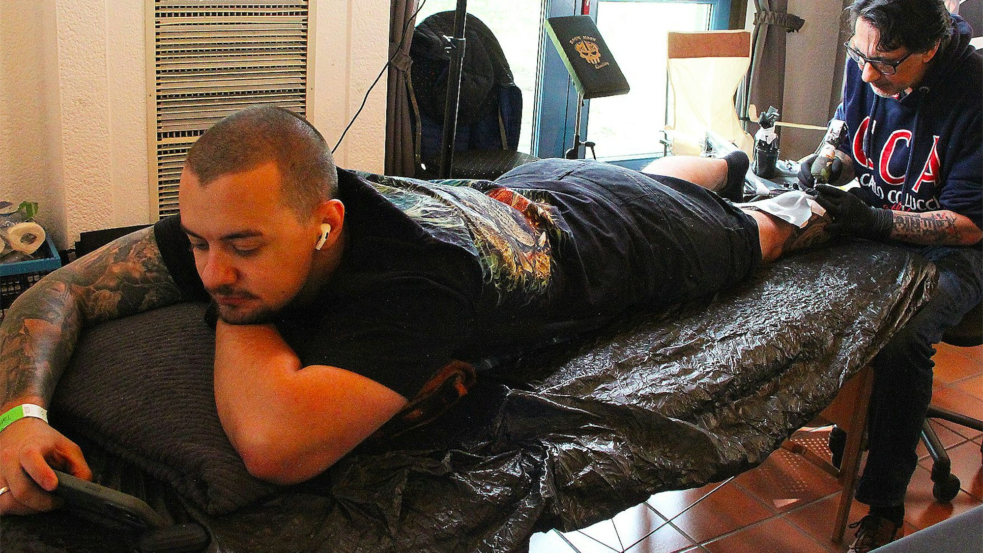 Ein Mann liegt auf einer Liege und schaut auf sein Smartphone, während ein anderer Mann seinen linken Unterschenkel tätowiert.