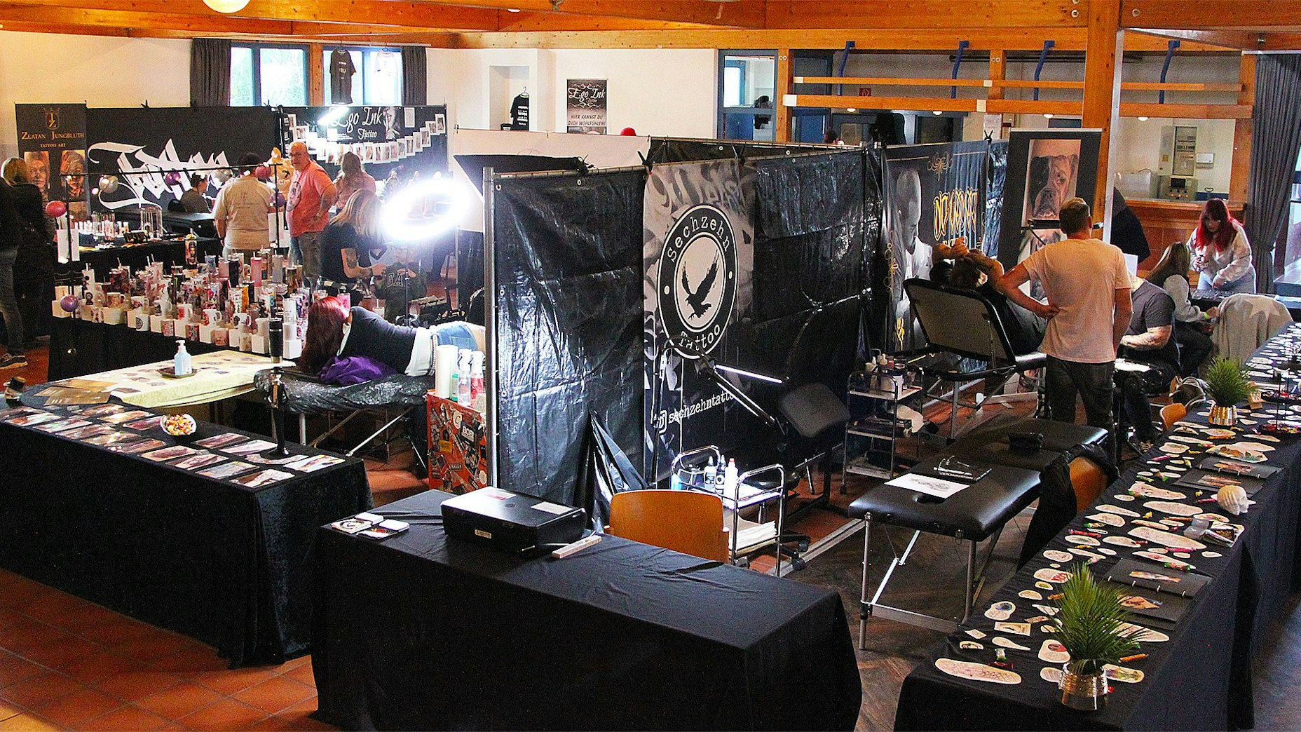 Mehrere schwarze Tische, Stellwände und Liegen stehen in einem Raum. Auf den Tischen liegen Tattoo-Motive.