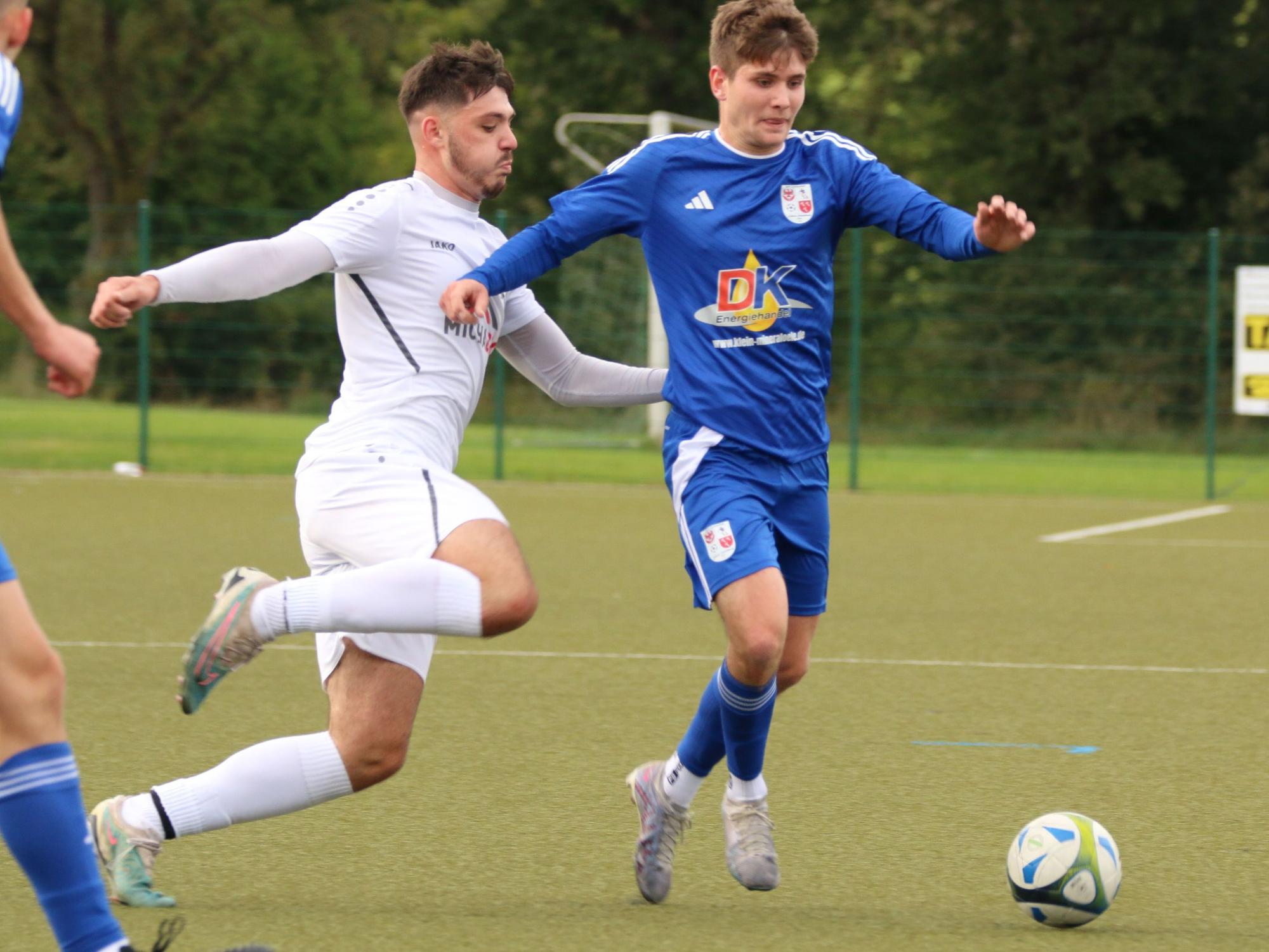 Ein Fußballer läuft mit einem Ball am Fuß, die Arme sind ausgestreckt, von hinten greift ein Gegenspieler an Es könnte weh tun.