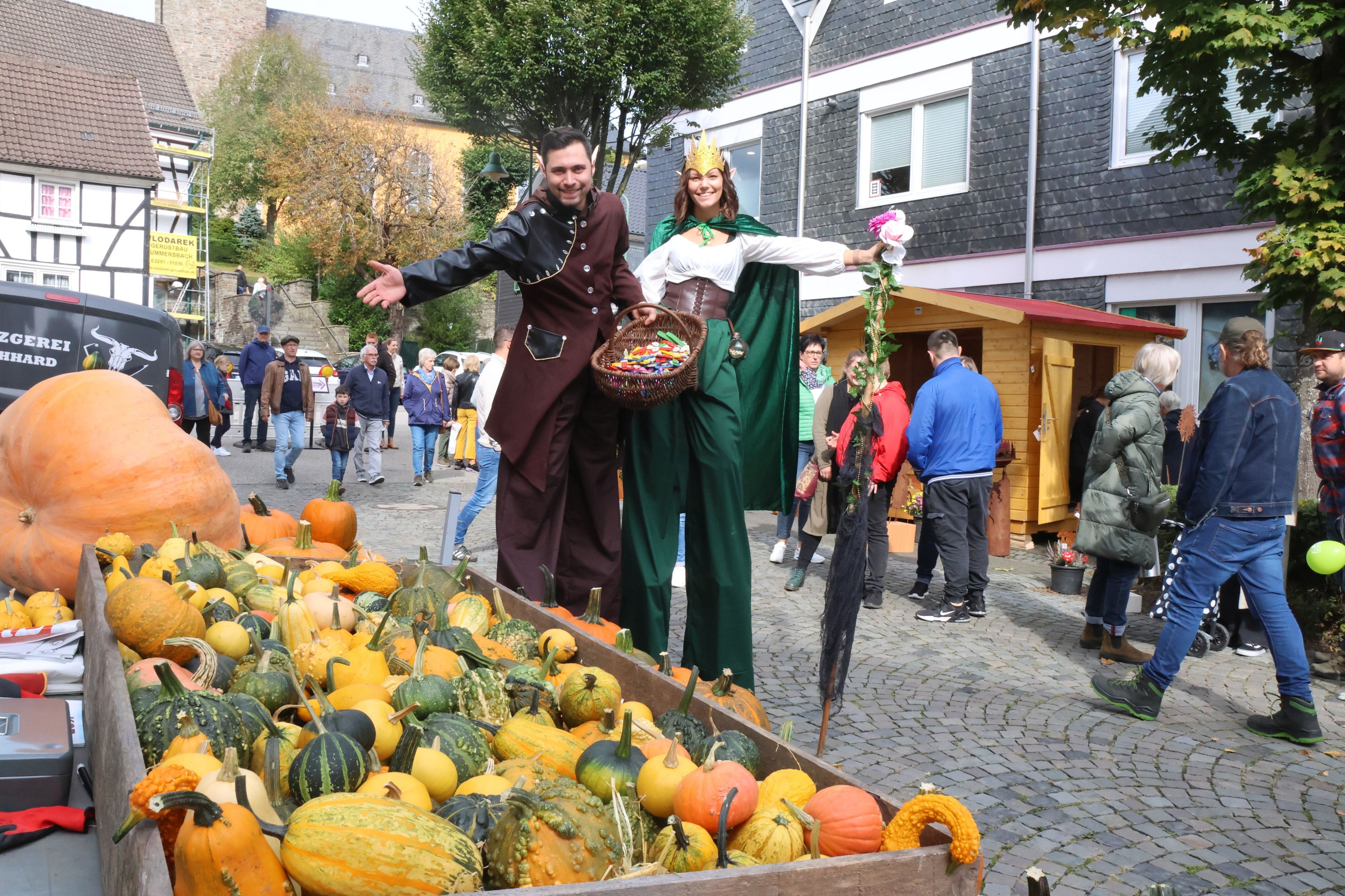 Zwei Personen auf Stelzen und im Waldelfenkostüm verteilen auf einem Erntedankfest Blumen und Süßigkeiten.