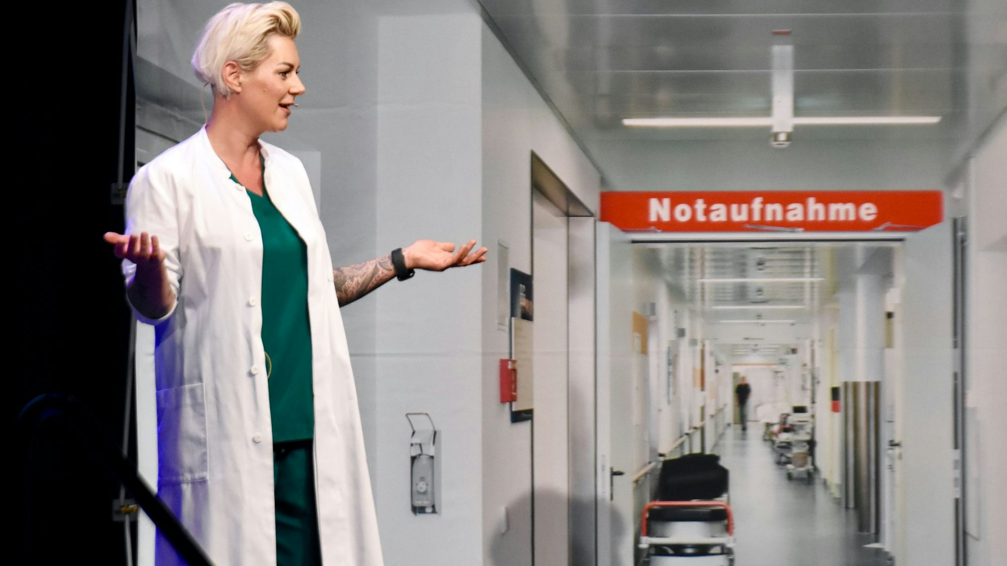 Carola Holzner alias Doc Caro, eine blonde Frau im Ärztekittel, steht in einr Notaufnahme.