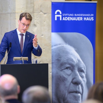 Ein Man steht an einem Pult, daneben befindet sich ein Aufsteller mit einem großen Porträt Konrad Adenauers.