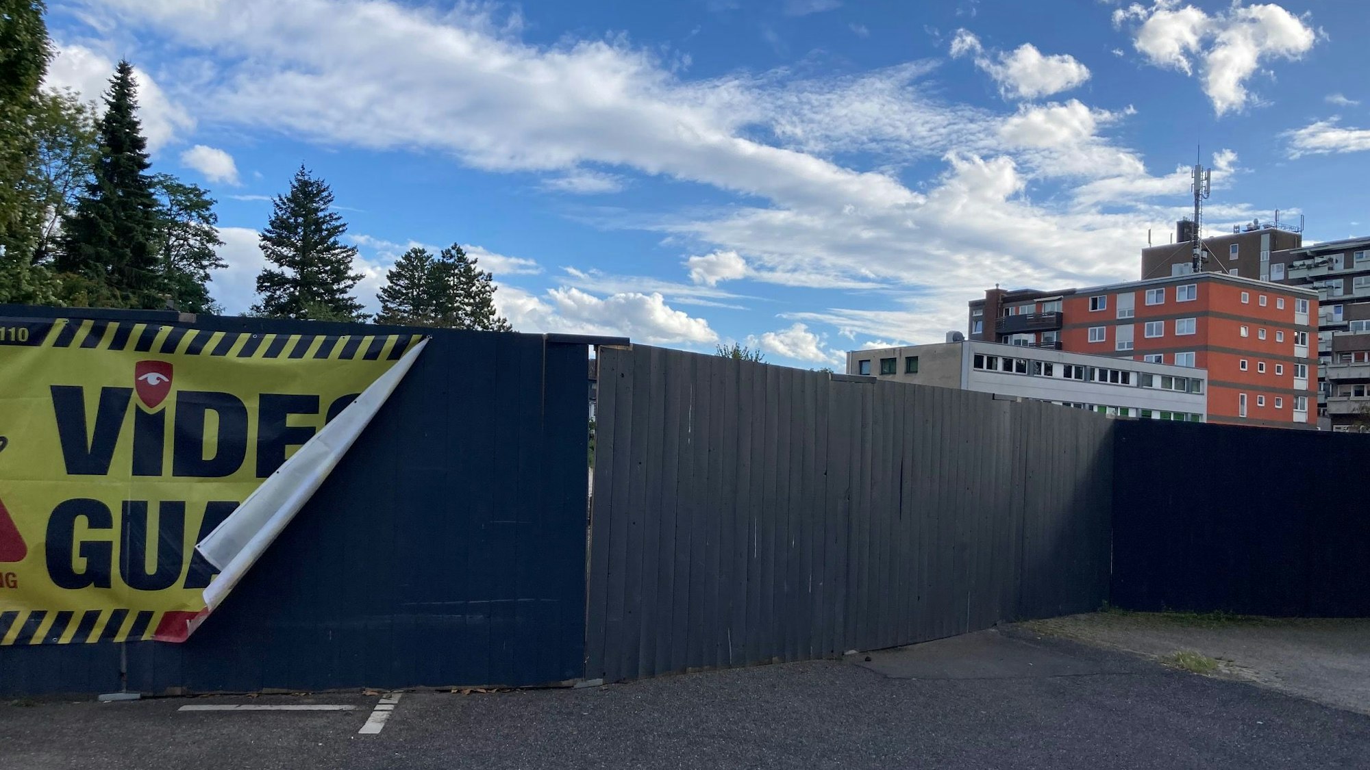 Ein hoher dunkler Bretterzaun ist zu sehen, an dem teilweise ein gelb-schwarzes Transparent mit der Aufschrift „Video Guard“ hängt.