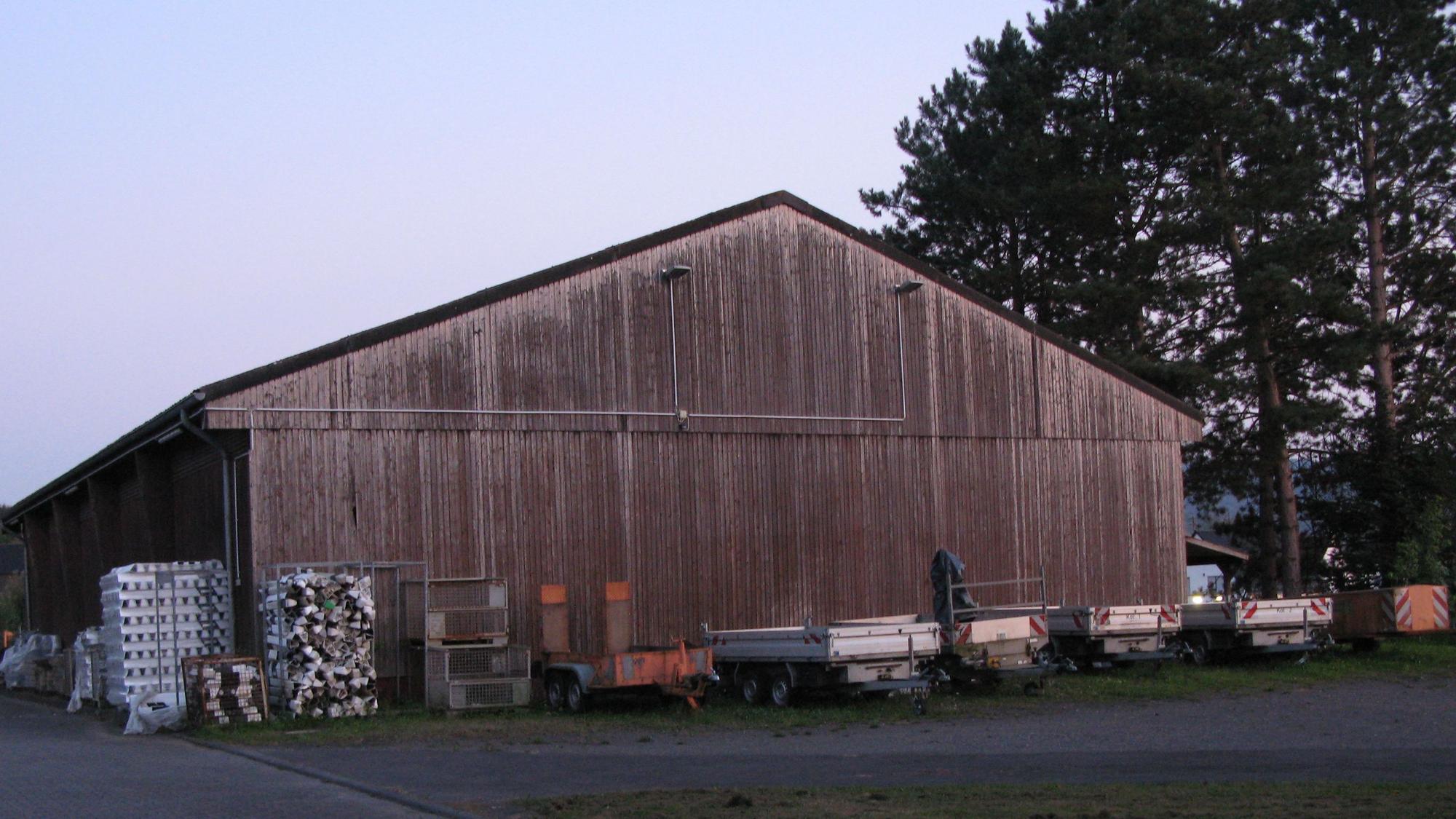 Eine alte Halle mit Satteldach und Holzlattenfassade, vor der einige Anhänger stehen.