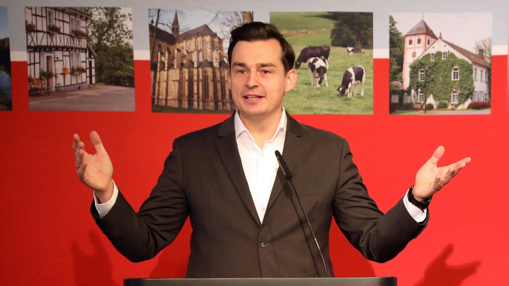 Der Bergisch Gladbacher Bürgermeisterkandidat von SPD und Grünen, Marcel Kreutz (SPD) spricht vor einer roten Wand mit Bildern aus Rhein-Berg.