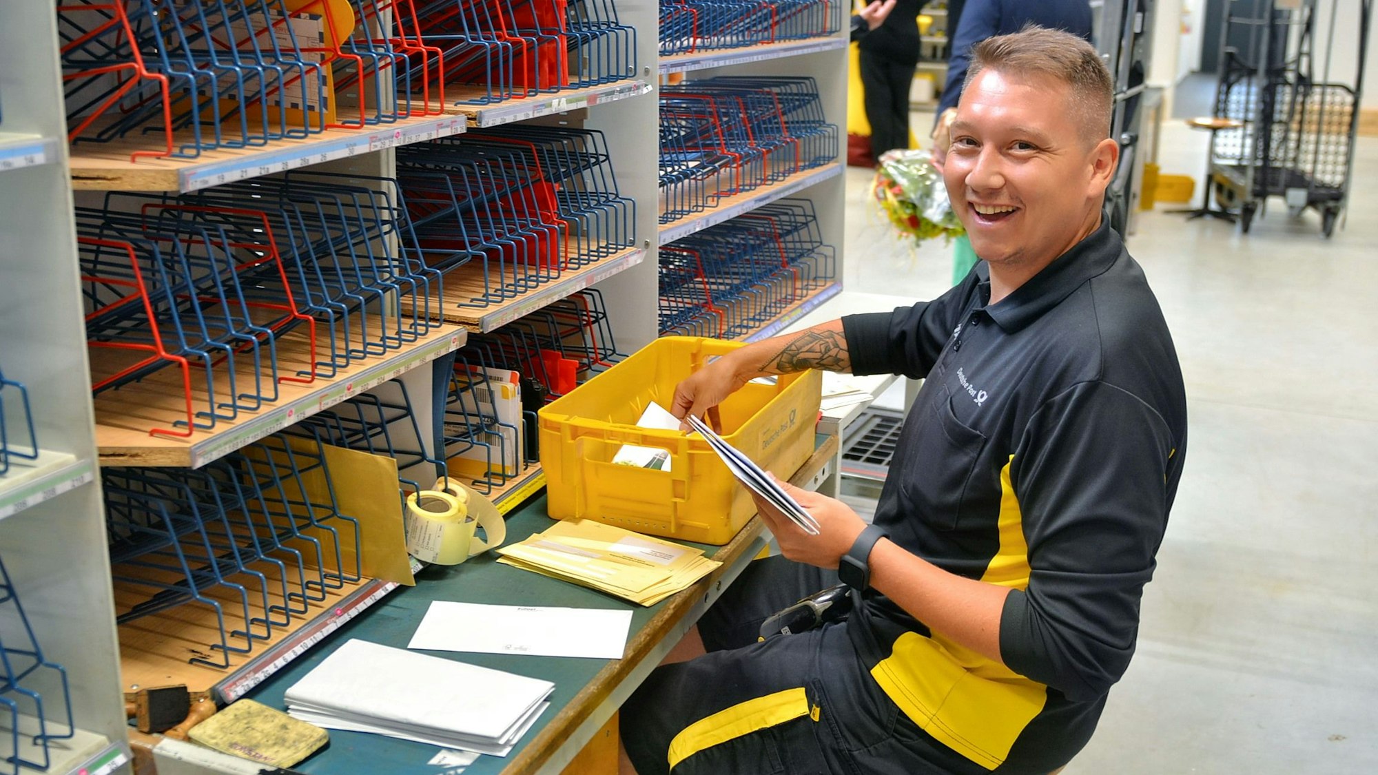 Kevin Söntgen trägt die schwarz-gelbe Dienstkleidung der Post. Er sitzt an einem Tisch vor einer Regalreihe und sortiert Briefe in eine Kiste.