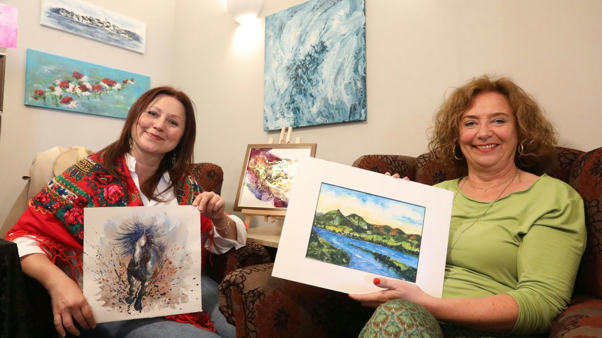 Zwei Frauen sitzen in Sesseln und zeigen ihre Kunstwerke, hinter ihnen hängen weitere Arbeiten an der Wand.
