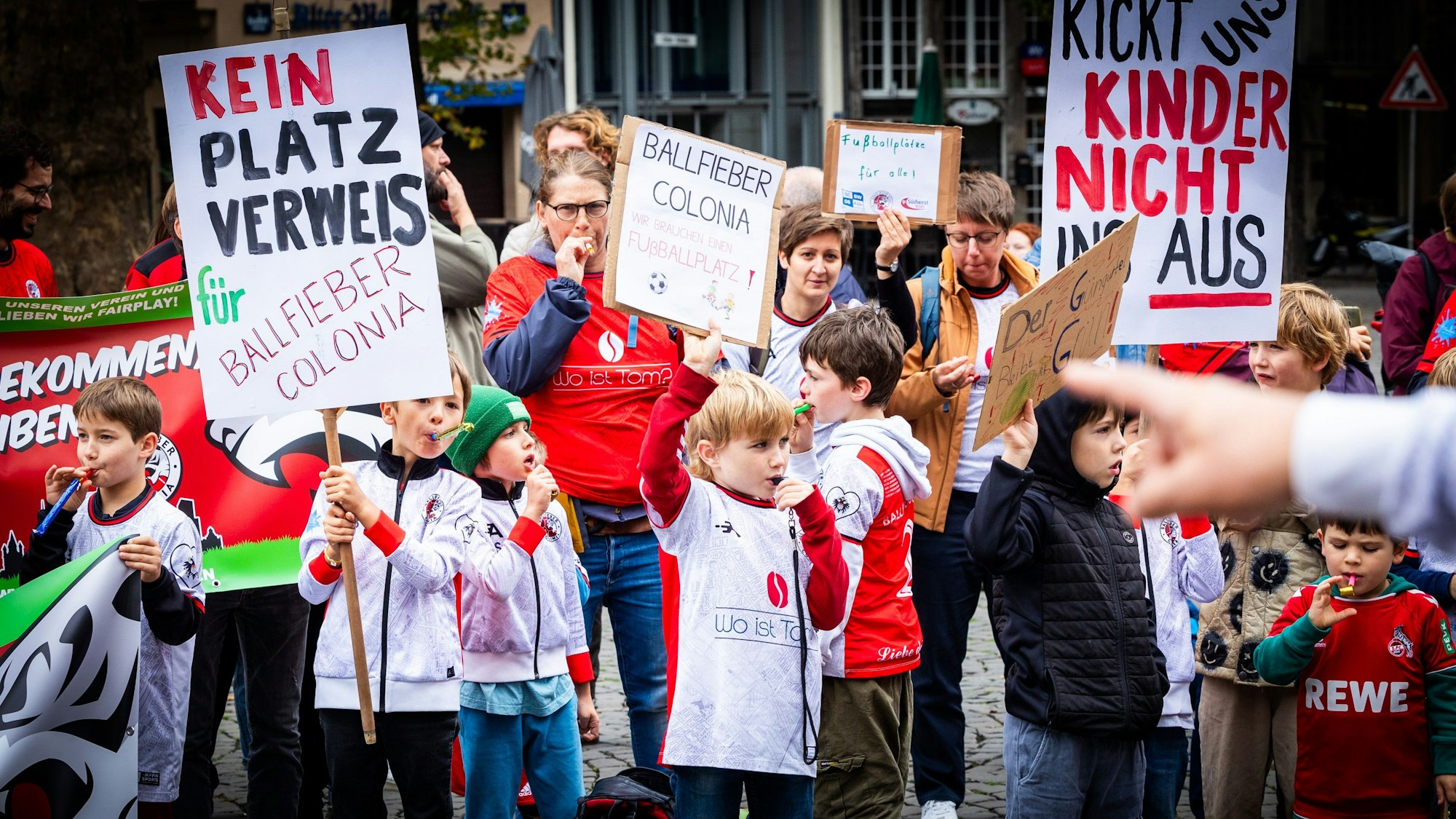 Kinder und Erwachsene halten bei einer Demo Plakate in den Händen und haben Trillerpfeifen im Mund.