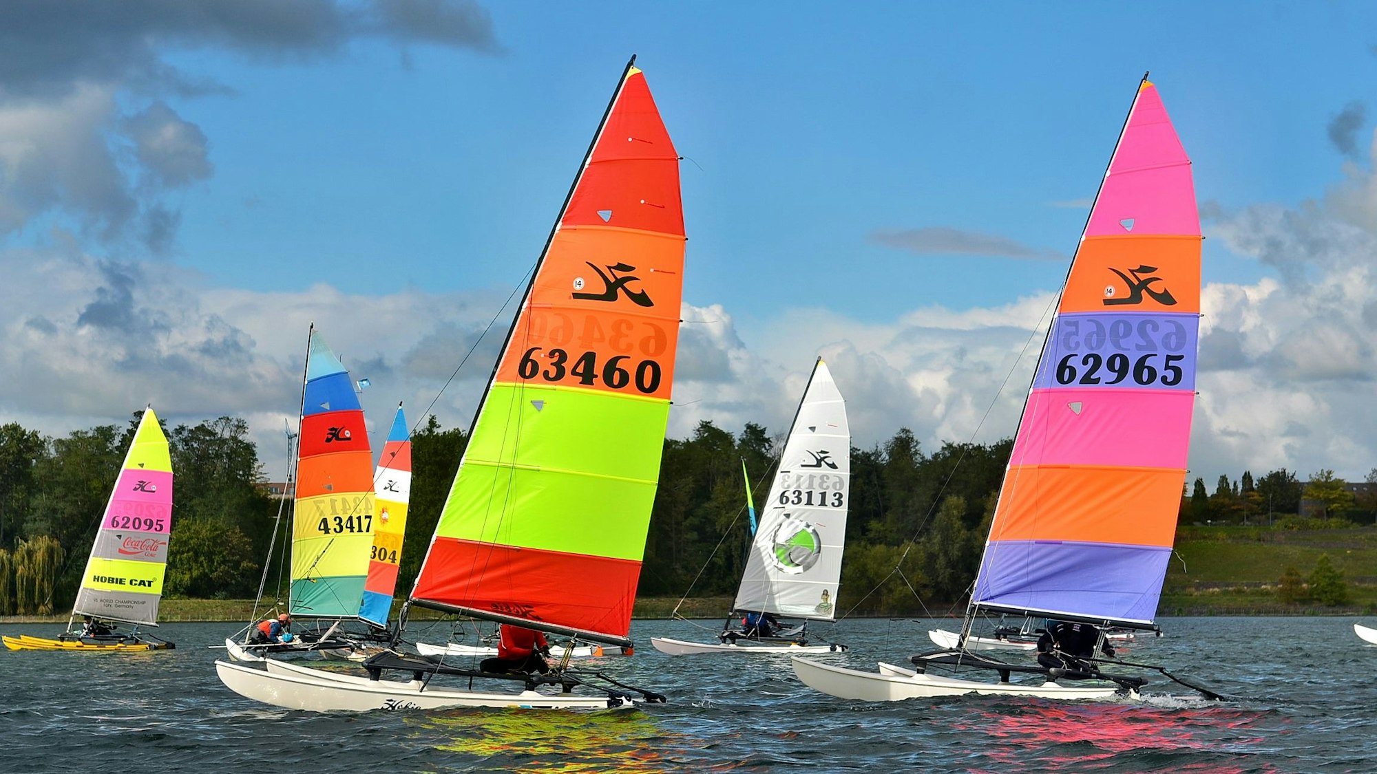 Sechs Boote mit bunten Segeln fahren auf dem Wassersportsee in Zülpich.