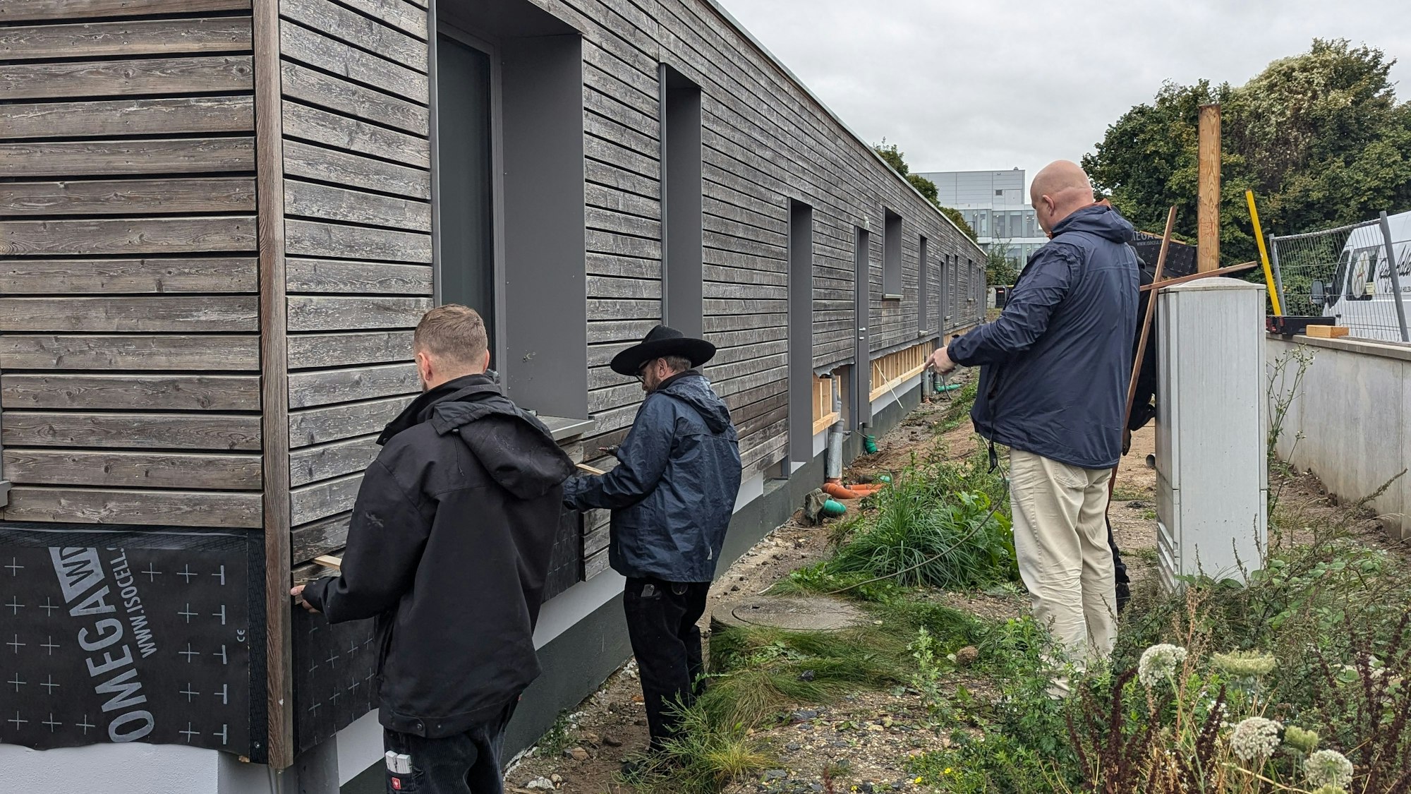 Das Foto zeigt Handwerker, die an der Holzfassade eines eingeschossigen Gebäudes arbeiten.