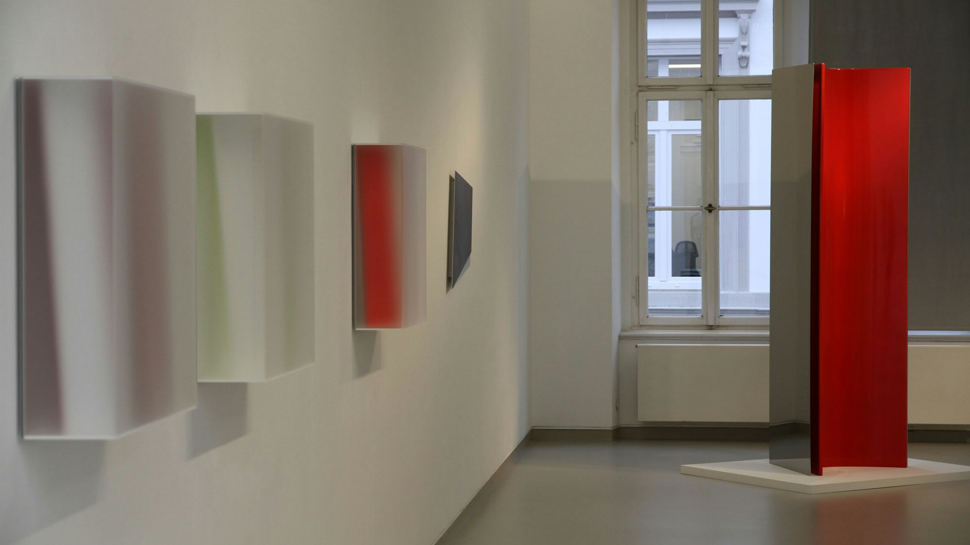 Quadratische Kunstobjekte an einer Wand,rechts eine Stele aus spiegelndem und rot lackiertem Metall