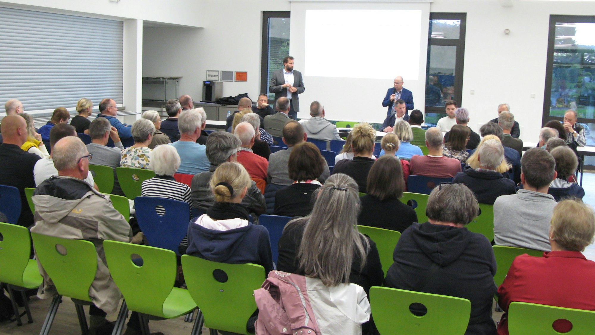 Rund 70 Menschen sitzen in sechs Reihen auf grünen Stühlen. Vorne stehen zwei Männer vor einer Leinwand.