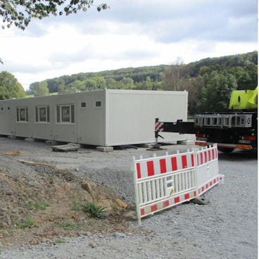 Eine Reihe hellgrauer Wohncontainer auf einem Schotteruntergrund, daneben eine Baustellenabsperrung.