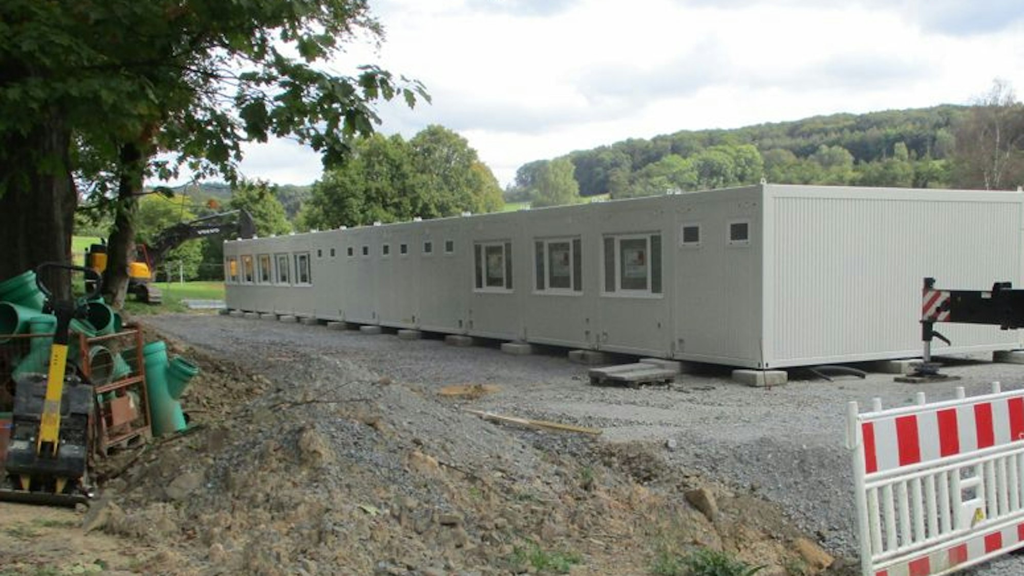 Eine Reihe hellgrauer Wohncontainer auf einem Schotteruntergrund, daneben eine Baustellenabsperrung.