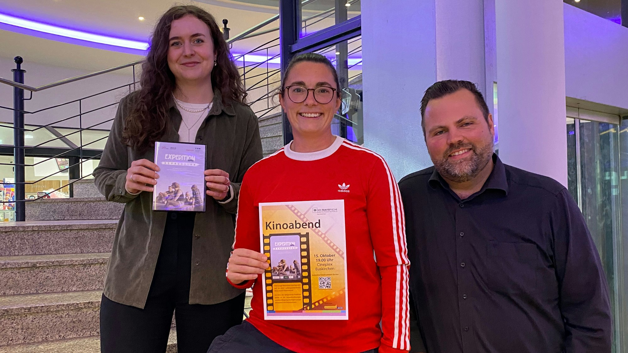 Zwei Frauen und ein Mann stehen auf der Treppe zum Cineplex und präsentieren die DVD-Hülle und ein Plakat zu der Filmvorführung.