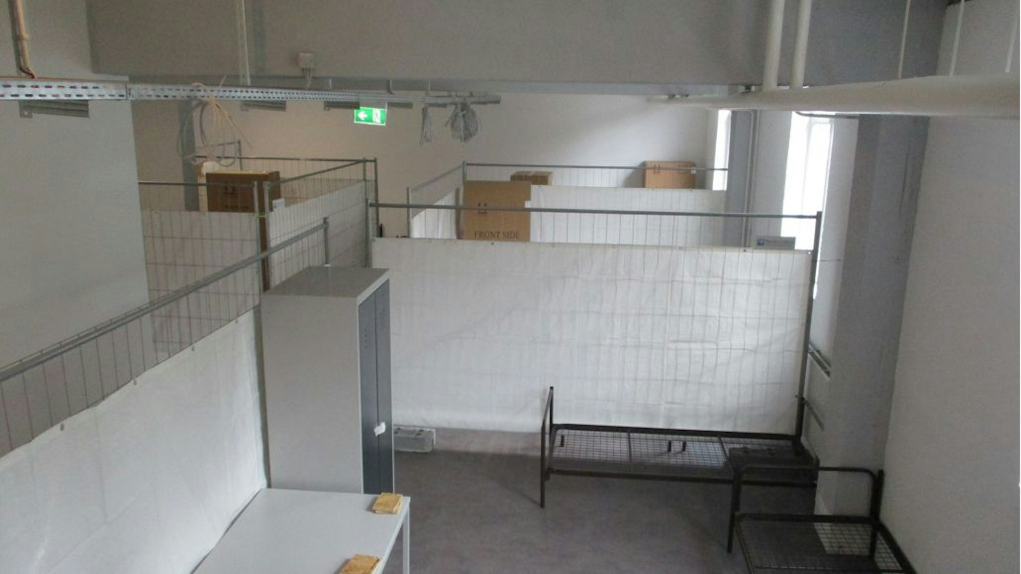 Blick in ein Zimmer der Notunterkunft, dünne Trennwände, ein Tisch, ein Schrank, ein Bett.