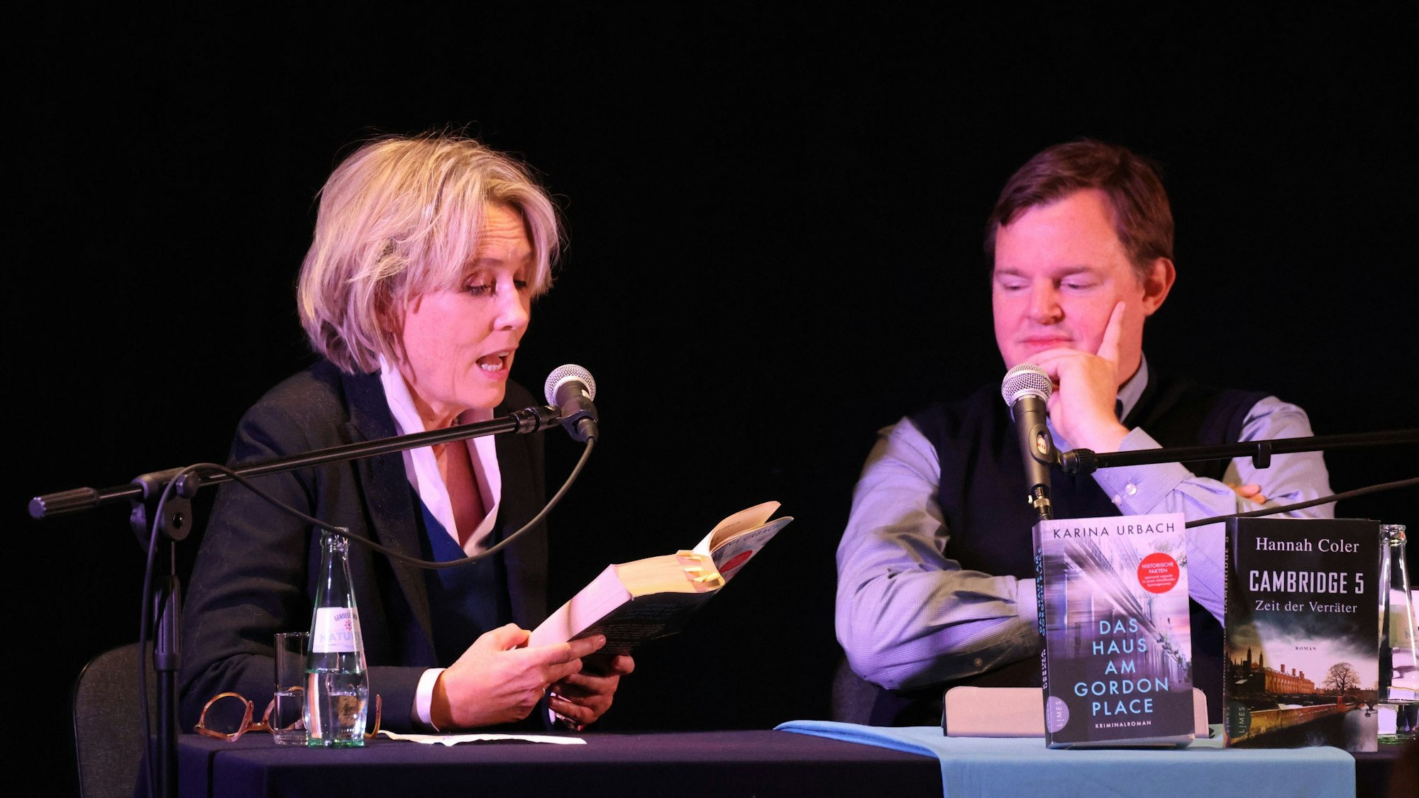 Karina Urbach und Mike Altwicker sitzen bei einer Lesung nebeneinander. Sie liest aus ihrem Buch vor.