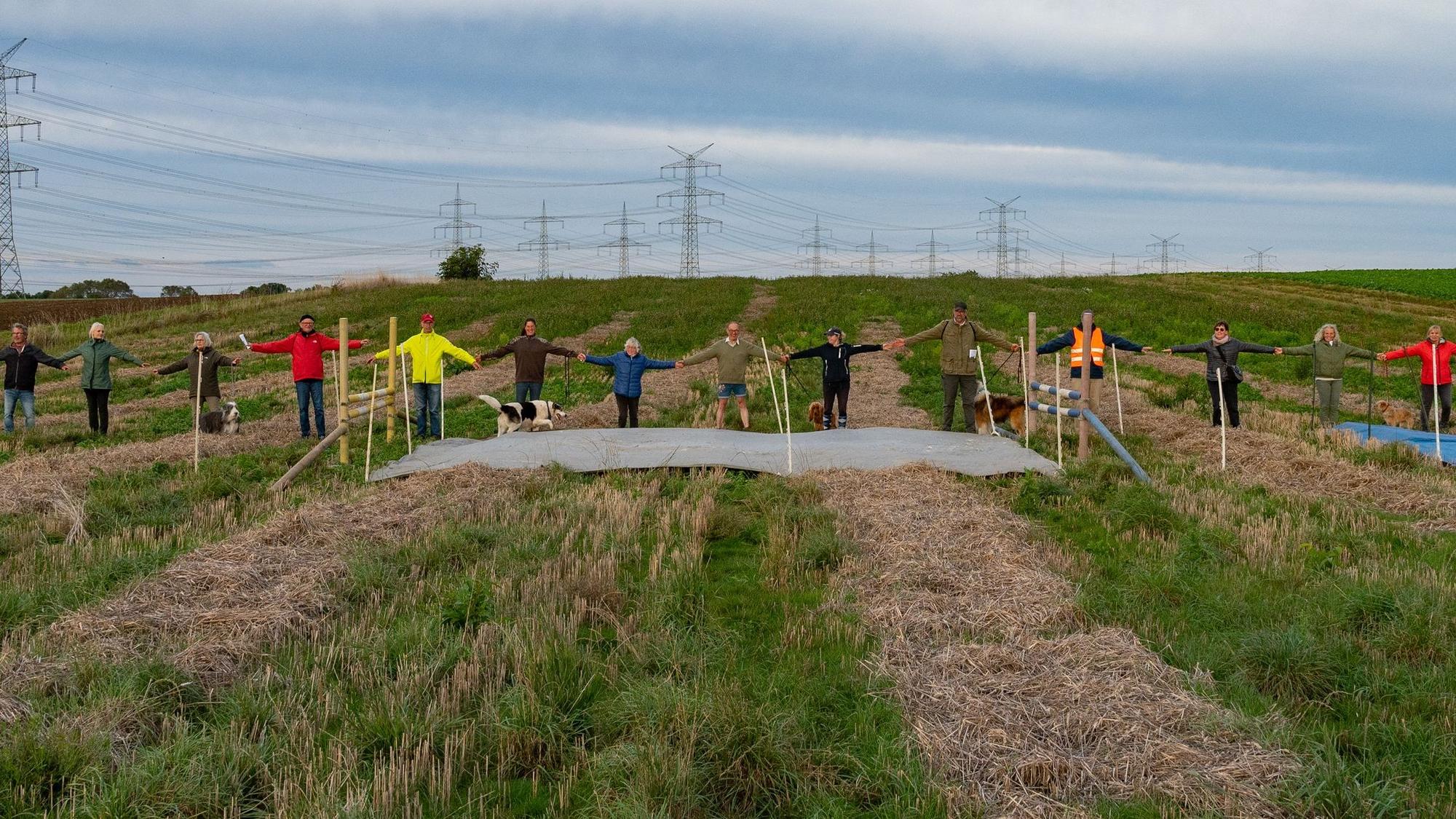 Menschen stehen auf einem Feld, sie halten sich an den Händen, um zu verdeutlichen, wie breit die geplante Landesstraße werden soll.