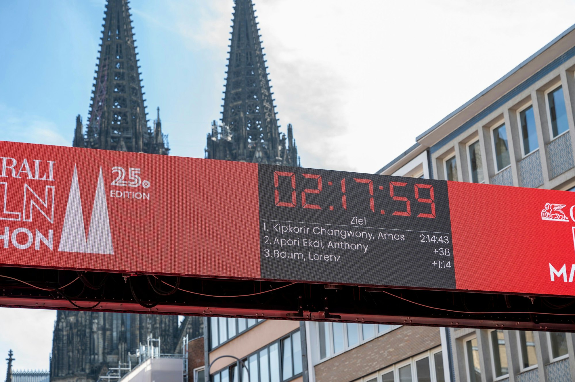 Eine Anzeigetafel zeigt die Zeit beim Köln-Marathon an. Dahinter ist der Dom zu sehen.