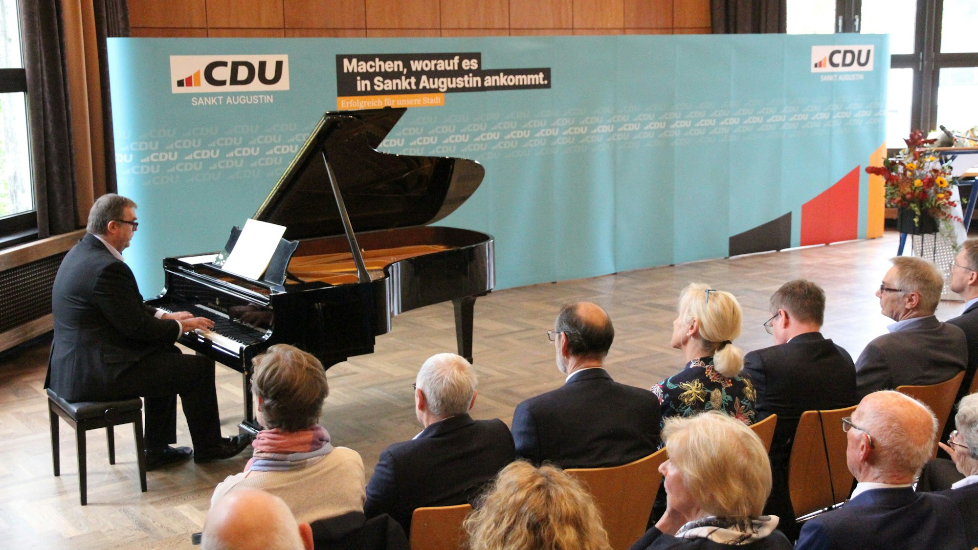 Ein Mann an einem Konzertflügel und Zuhörer auf Stühlen in einem Saal