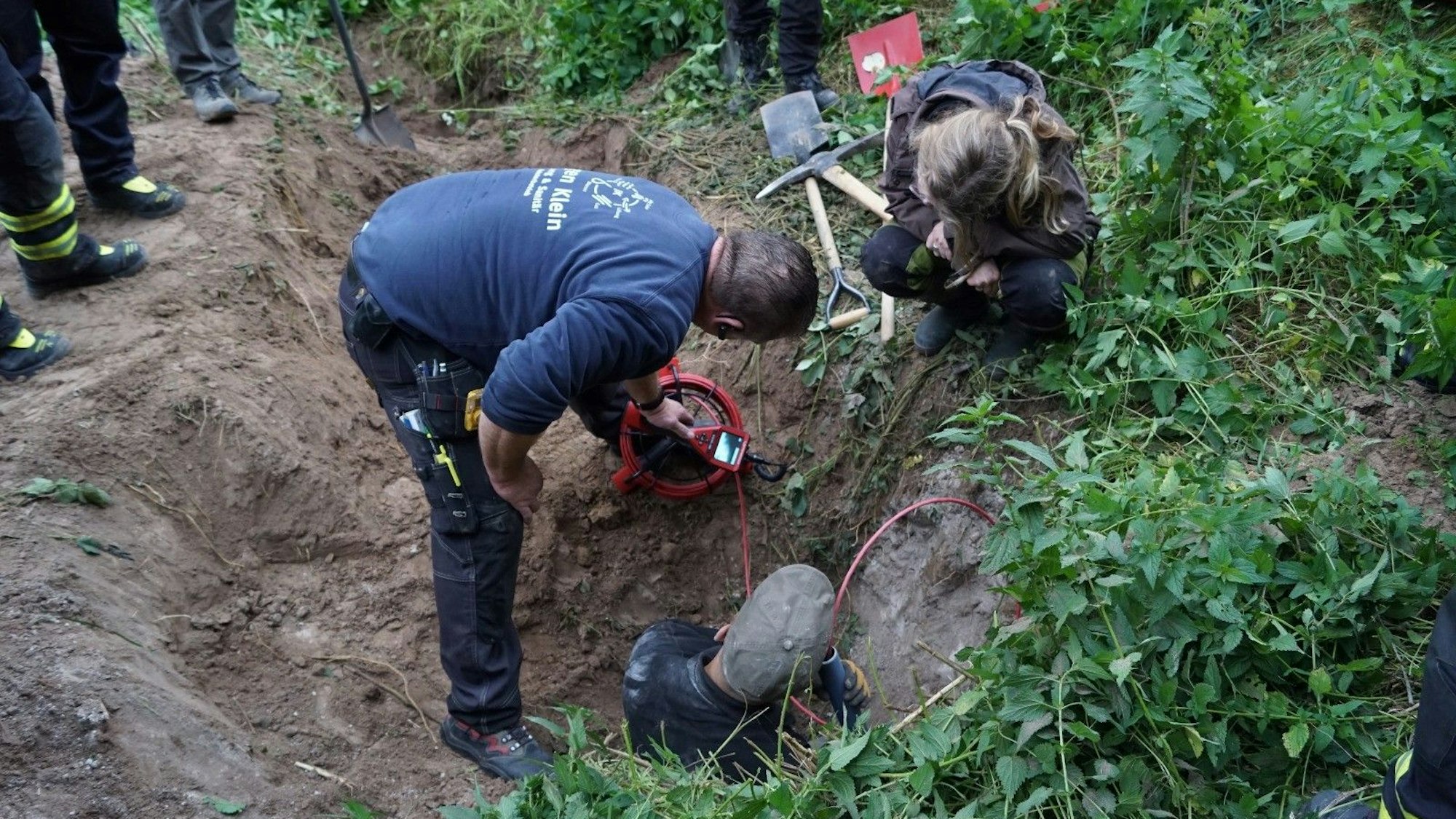 Ein Mann steht in einem Dachsbau. Ein weiterer Mann und eine Frau beugen sich vor und schauen auf ein Display einer Spezialkamera.