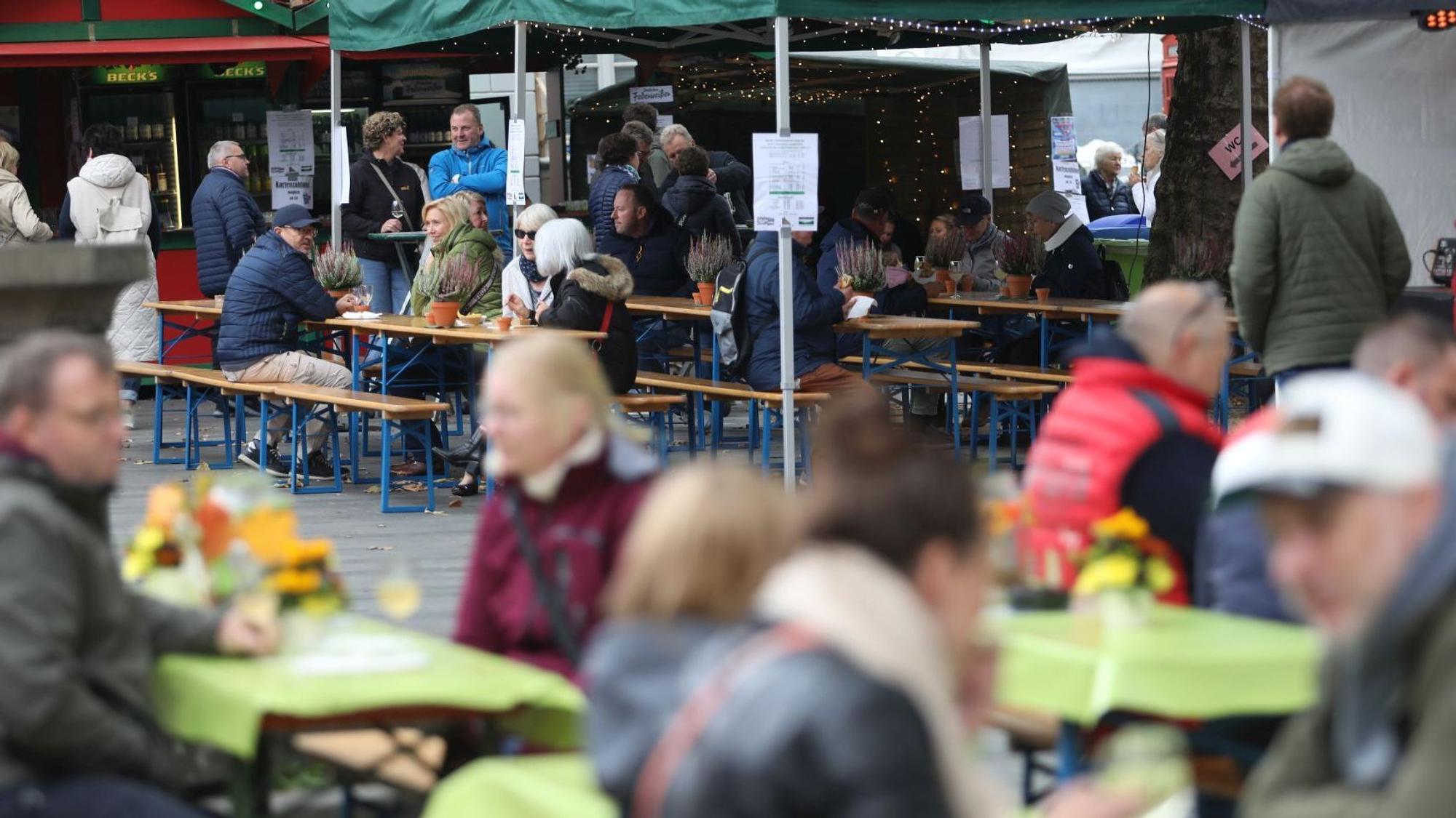 Menschen sitzen an Tischen auf dem Marktplatz, im Hintergrund stehen zwei Weinstände.