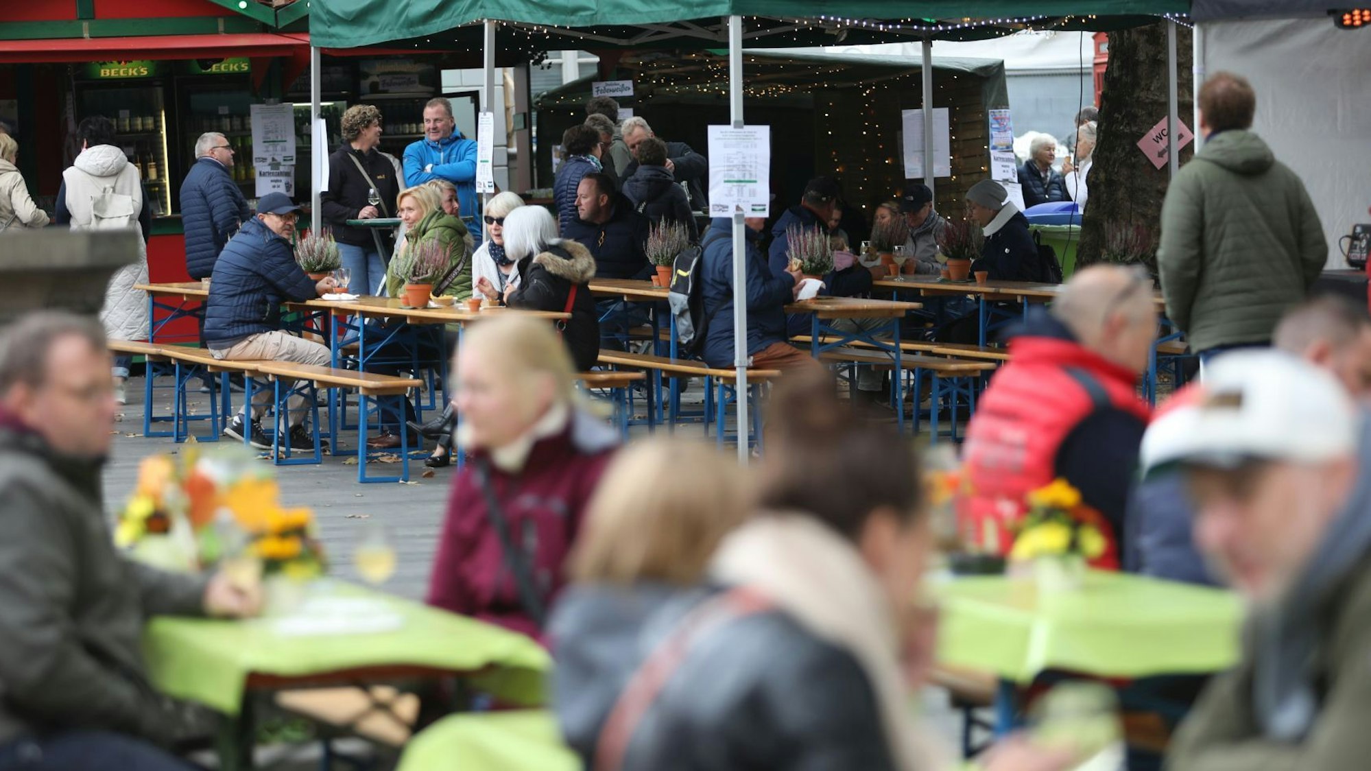 Menschen sitzen an Tischen auf dem Marktplatz, im Hintergrund stehen zwei Weinstände.
