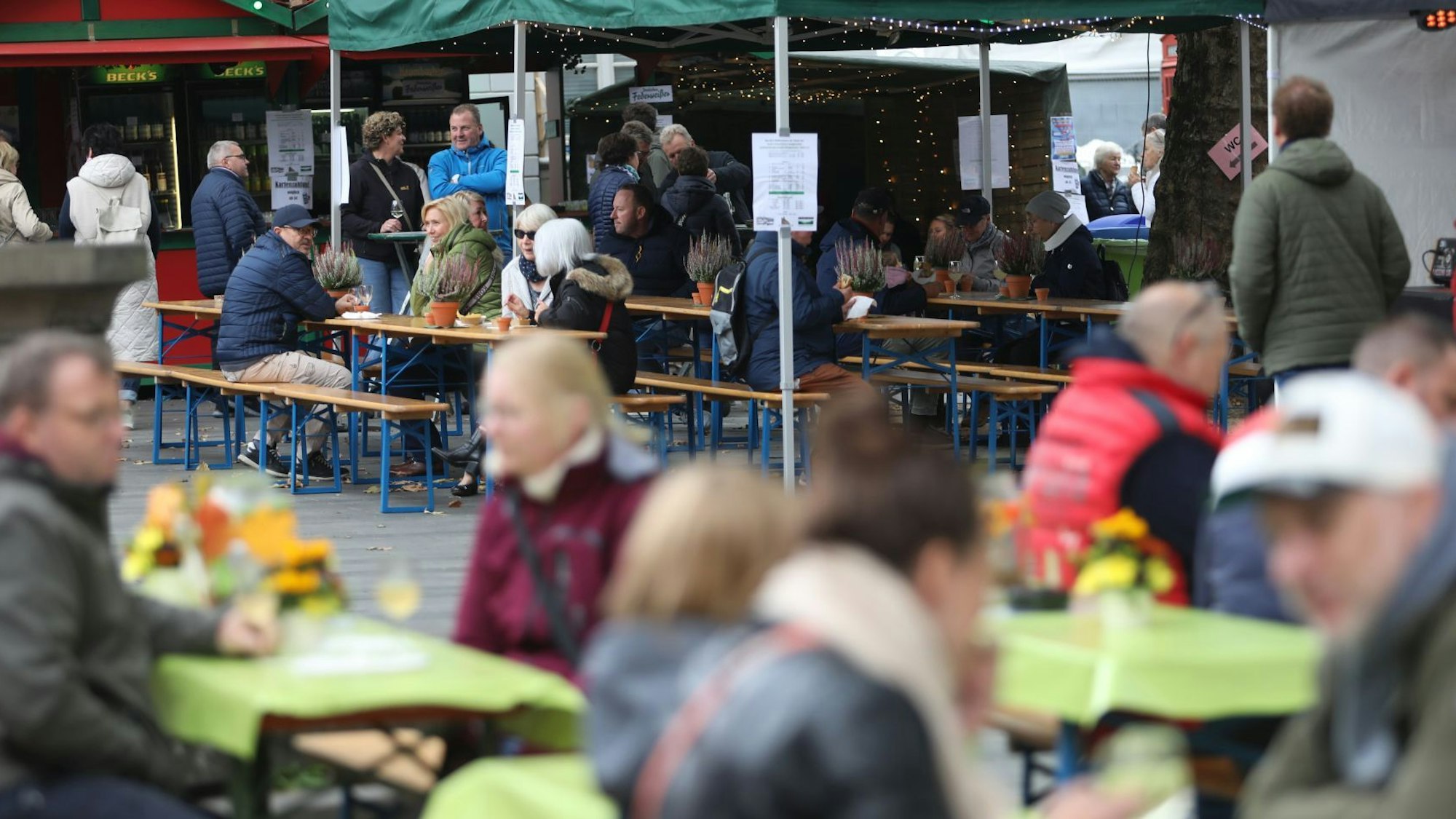 Menschen sitzen an Tischen auf dem Marktplatz, im Hintergrund stehen zwei Weinstände.