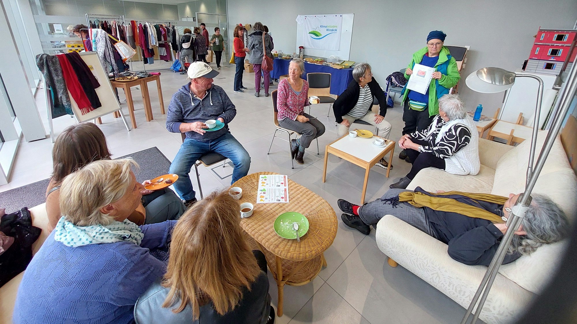 Menschen sitzen auf Stühlen und einer Couch im Kreis, im Hintergrund stehen Kleiderständer.