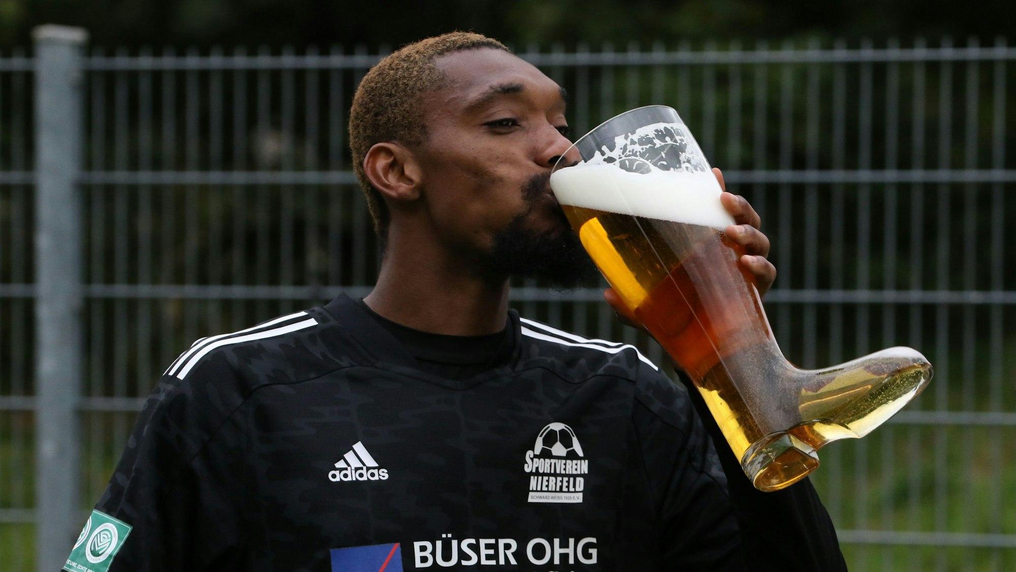 Fußballer Covenant Smart vom SV Niefeld hat mit seiner Mannschaft gewonnen und trinkt einen Schluck Bier aus einem großen Glas, das die Form eines Stiefels hat.