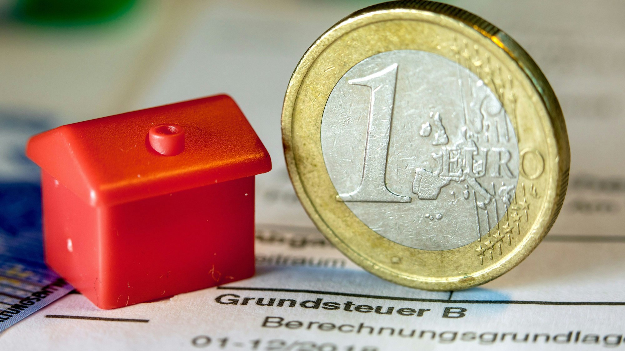 Eine Euro-Geldmünze sowie ein Spielzeughaus stehen auf einem Abgabenbescheid für die Entrichtung der Grundsteuer.