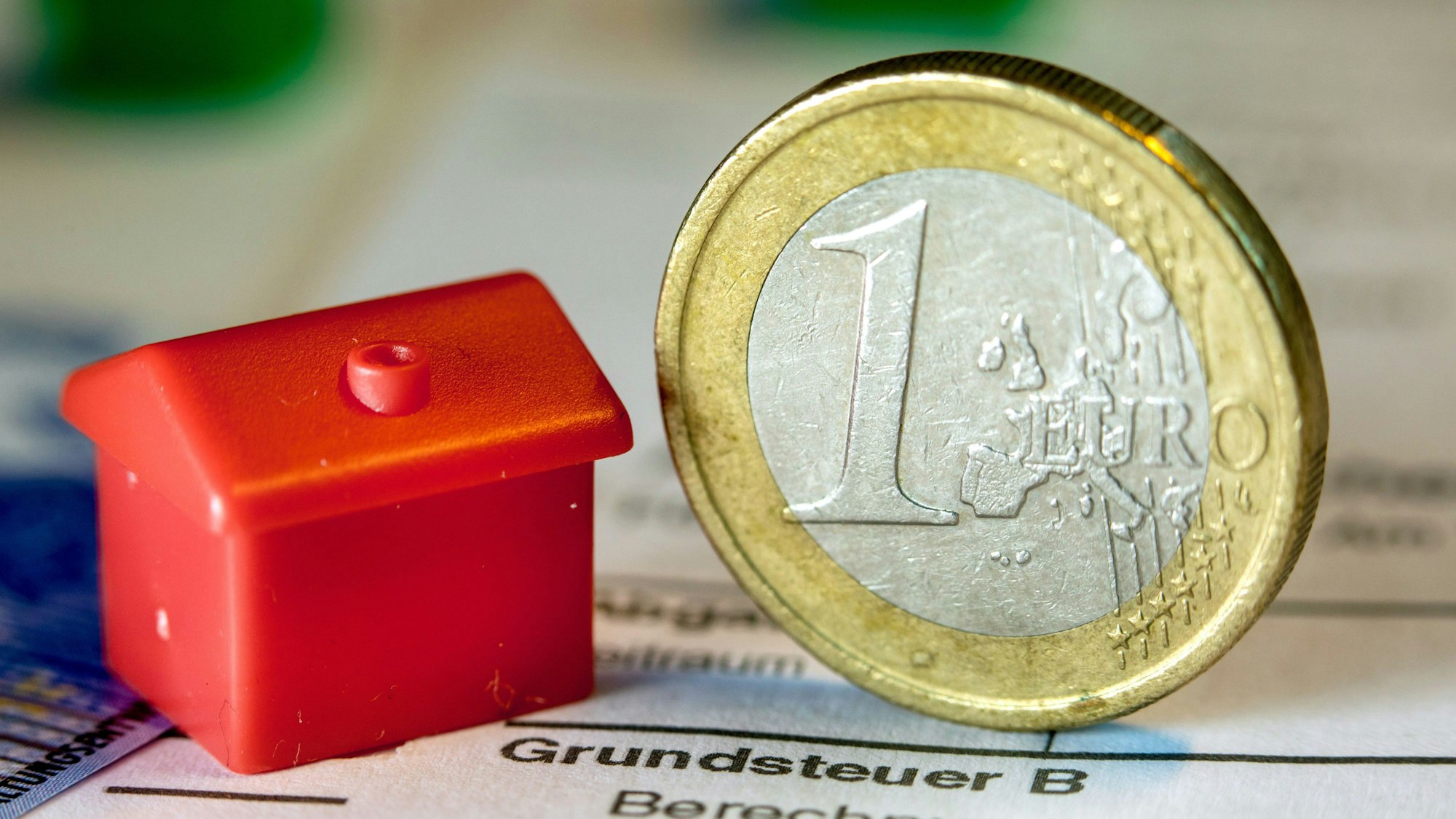 Eine Euro-Geldmünze sowie ein Spielzeughaus stehen auf einem Abgabenbescheid für die Entrichtung der Grundsteuer.