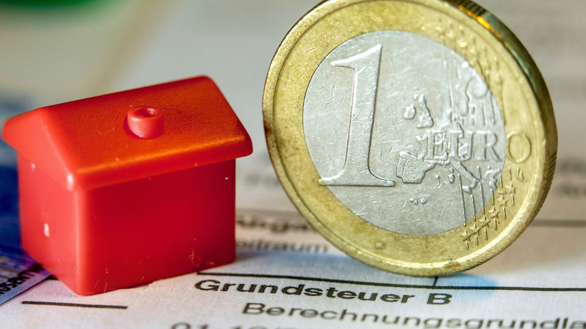 Eine Euro-Geldmünze sowie ein Spielzeughaus stehen auf einem Abgabenbescheid für die Entrichtung der Grundsteuer.