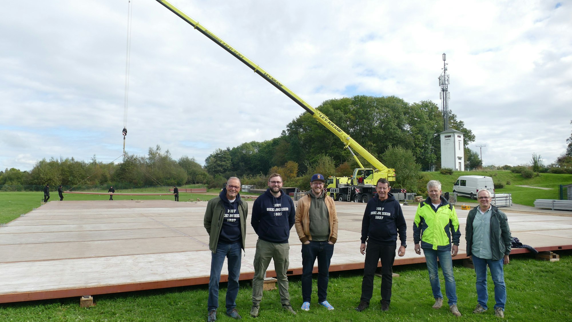 Sechs Männer stehen vor einer Fläche aus Platten, im Hintergrund ein Autokran.