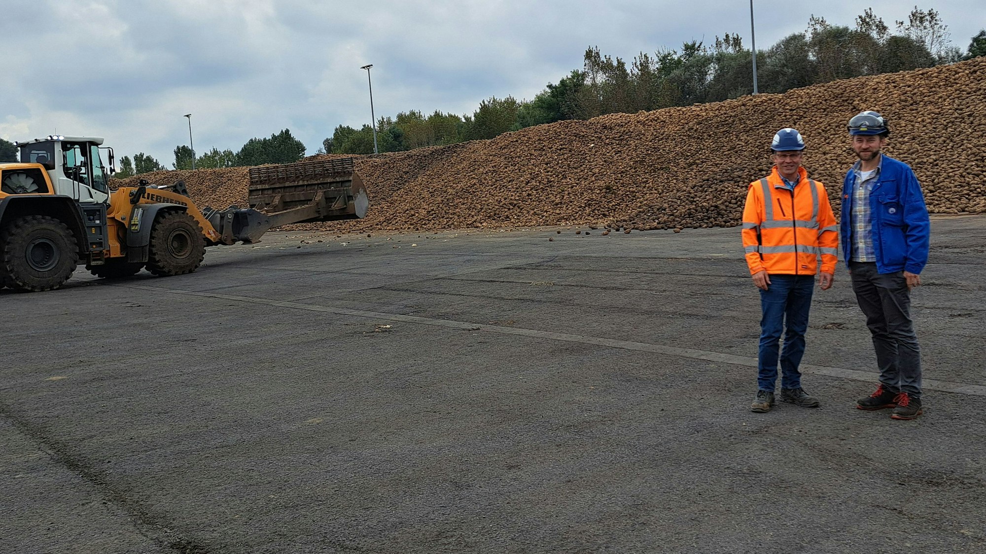 Andreas Gehlen (l.) und Markus Coenen stehen vor einem langgestreckten Berg Zuckerrüben. Links ist ein gelb-weißer Bagger zu sehen.