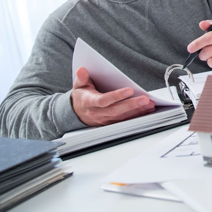 Ein Mann sitzt mit einem dicken Aktenordner an einem Tisch, auf dem das Modell eines Wohnhauses steht.
