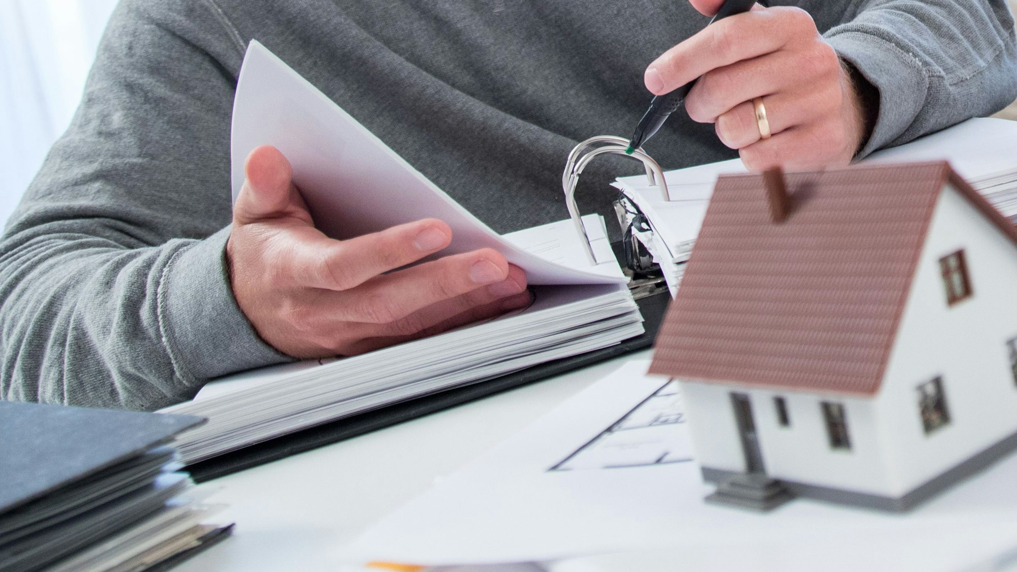 Ein Mann sitzt mit einem dicken Aktenordner an einem Tisch, auf dem das Modell eines Wohnhauses steht.